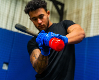 A baseball player in blue gloves grips a bat outfitted with the red Hitting Knob Bat Weight by Hitting Knob, preparing to swing in an indoor training facility.