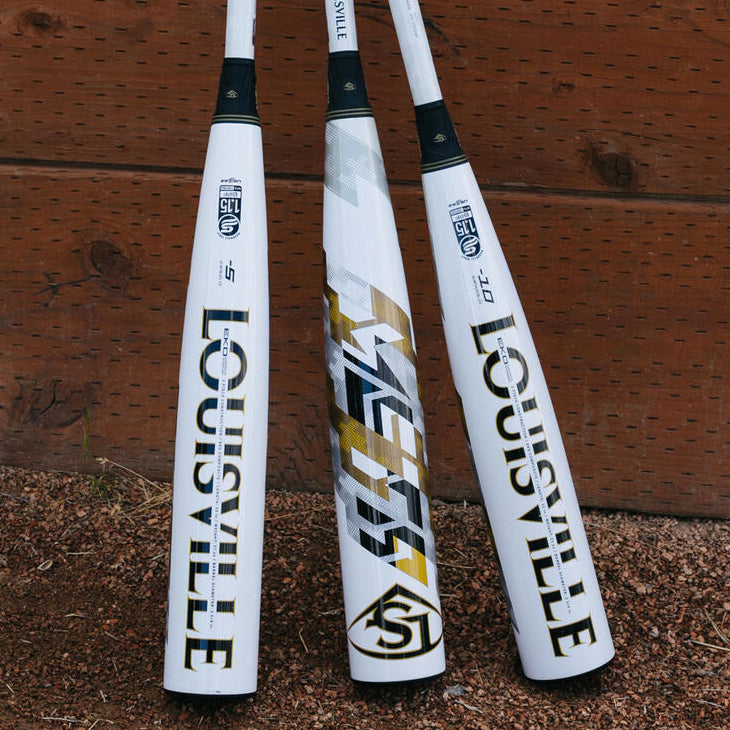 Three white Louisville Slugger baseball bats with gold and black accents stand upright on gravel against a wooden background.