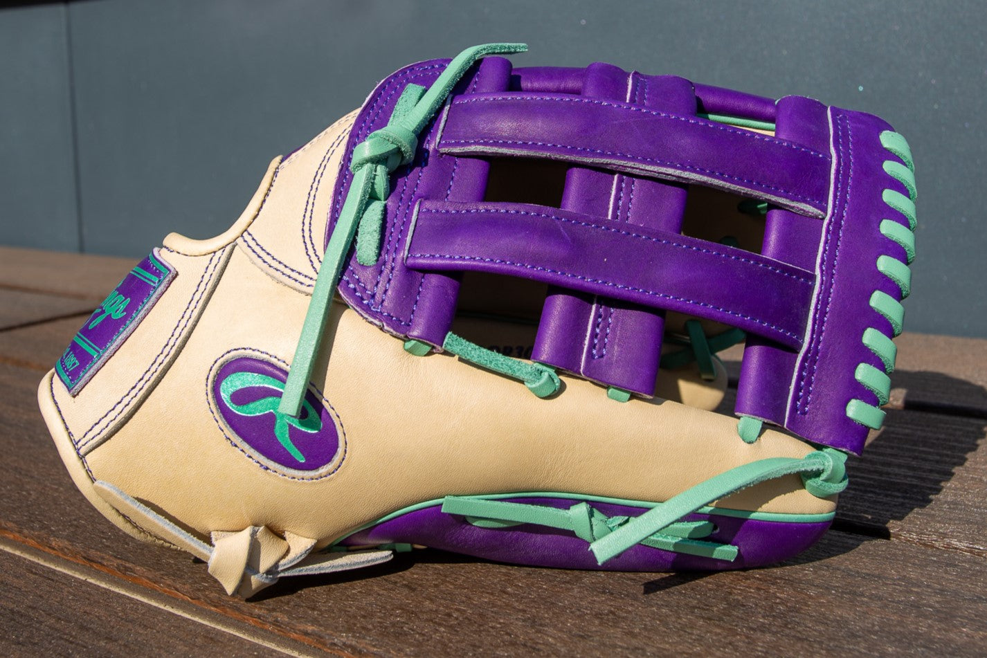 A tan Rawlings Heart of the Hide 12.75" Color Sync Glove (PROR3039-6CPU) with teal laces, purple trim, and a purple logo patch rests on a wooden surface—blending classic style with bold color accents.