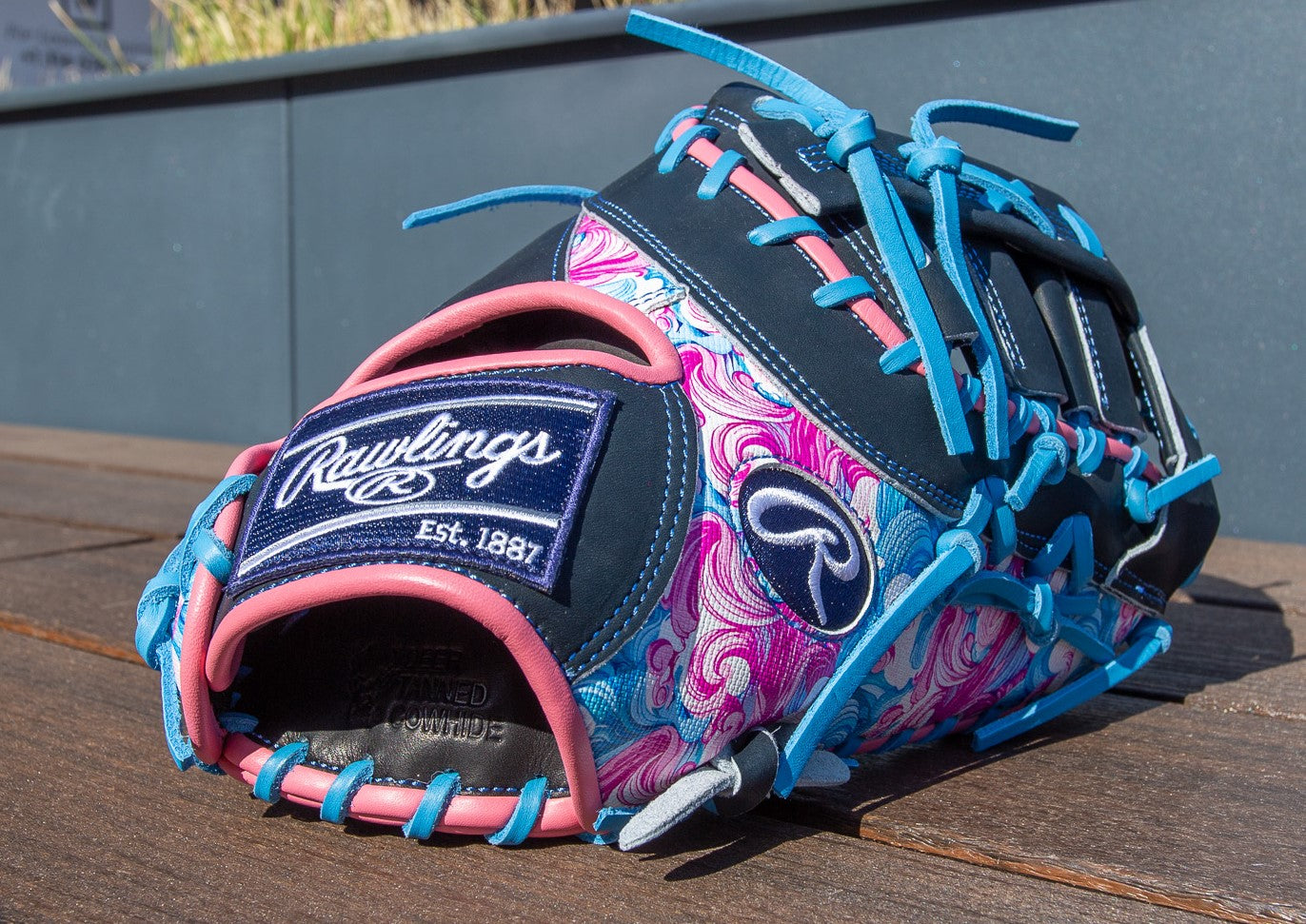 A vibrant Rawlings Heart of the Hide 13" Color Sync First Base Mitt (PRORDCT-10NSS) with a blue, pink, and purple swirl design, bright blue laces, and logo patch is shown outdoors on a wooden surface.