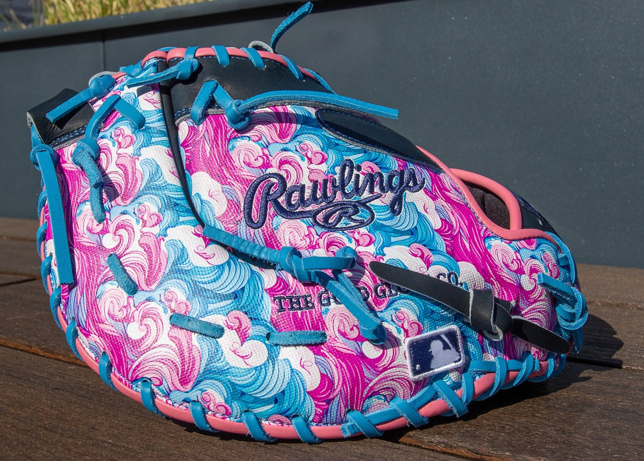 A Rawlings Heart of the Hide 13" Color Sync Baseball First Base Mitt (PRORDCT-10NSS) with a swirling pink, blue, and white pattern, blue stitching, and pink trim rests on wood. The MLB logo is visible on the glove.