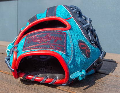 A Rawlings Heart of the Hide 11.5" Color Sync Baseball Glove (PROR204-2TE) in blue and black with red accents rests on a wooden surface, showcasing detailed stitching and embossed patterns.