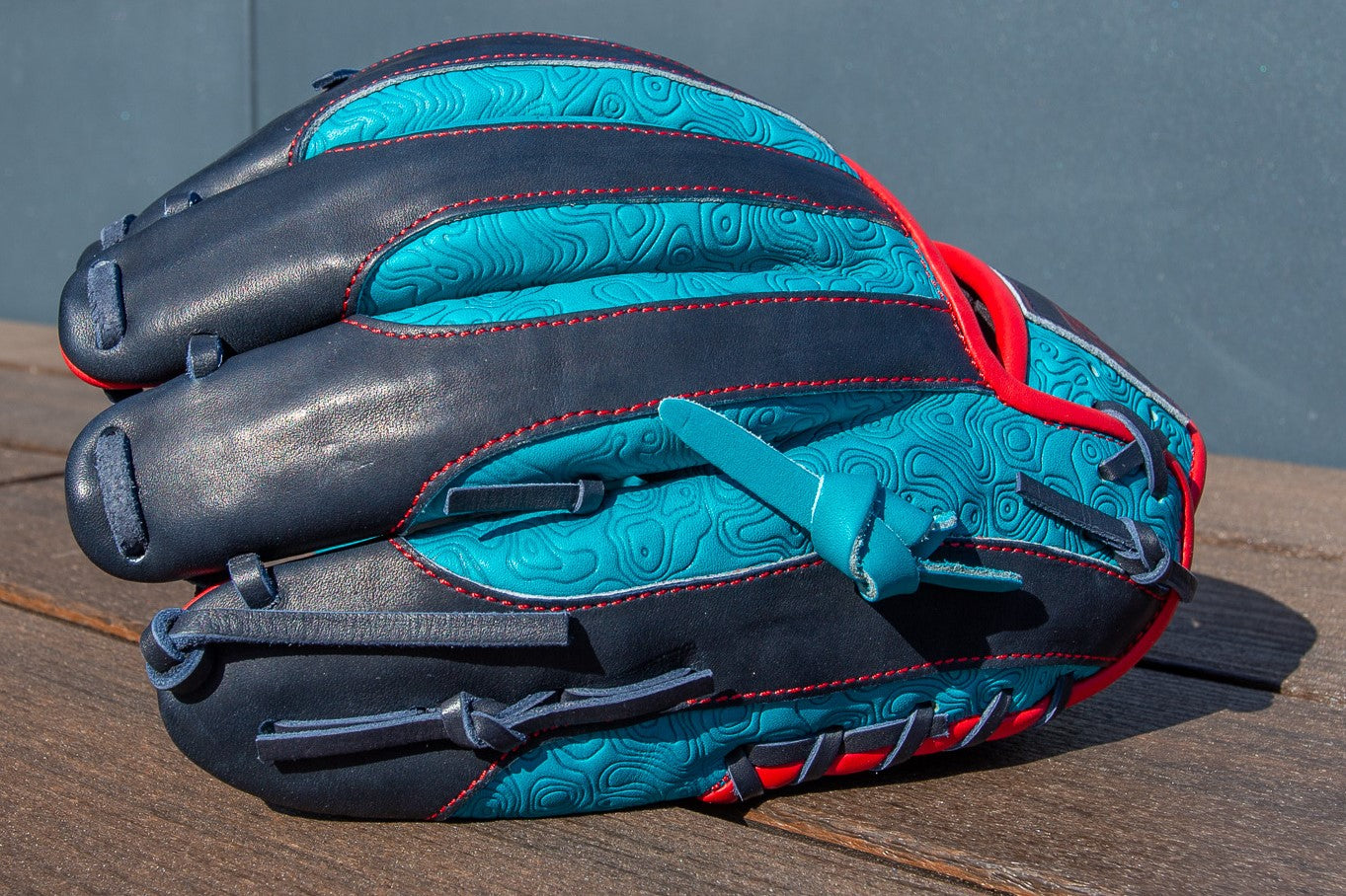 The Rawlings Heart of the Hide 11.5" Color Sync Baseball Glove (PROR204-2TE) boasts blue and black leather with red stitching and a topographic pattern, making a bold statement on the diamond as it rests on a wooden surface.
