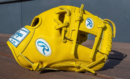 The Rawlings Heart of the Hide 11.5" Color Sync Baseball Glove (PROR204-2Y) features bright yellow leather with white and blue patches, displayed on a wooden surface in sunlight against a dark gray background.