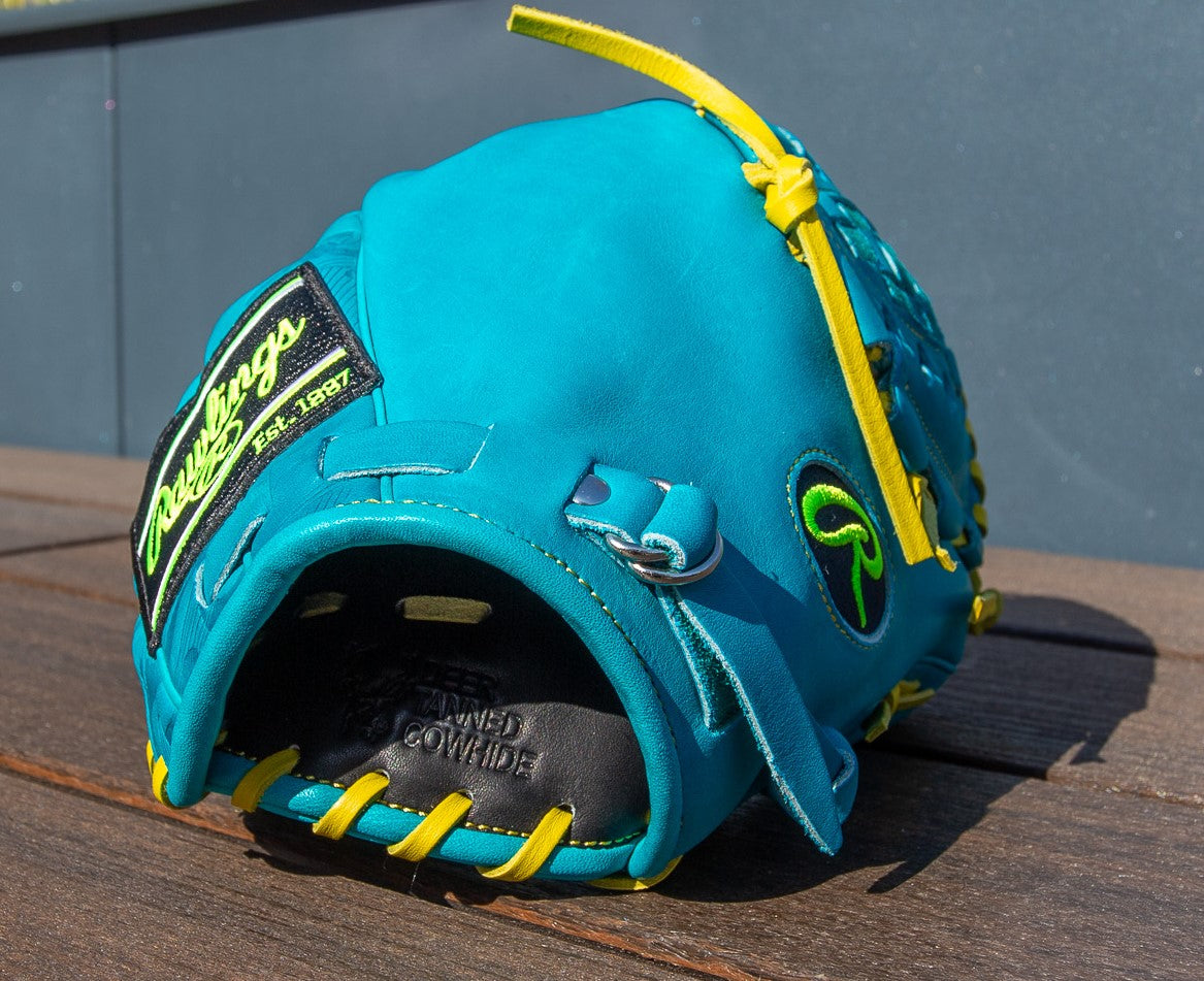 A Rawlings Heart of the Hide 12" Color Sync Baseball Glove (PROR206CFB-11TE) with teal leather, yellow laces, green and yellow logos, and black inner lining rests on a wooden surface in bright sunlight.