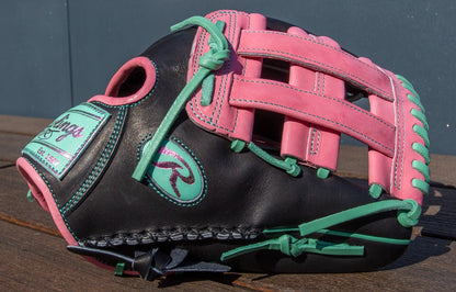 A Rawlings Heart of the Hide 12.25" Color Sync Baseball Glove (PROR207-6BP) featuring a black base, pink webbing, and teal-green laces rests on a wooden surface with a blue background.