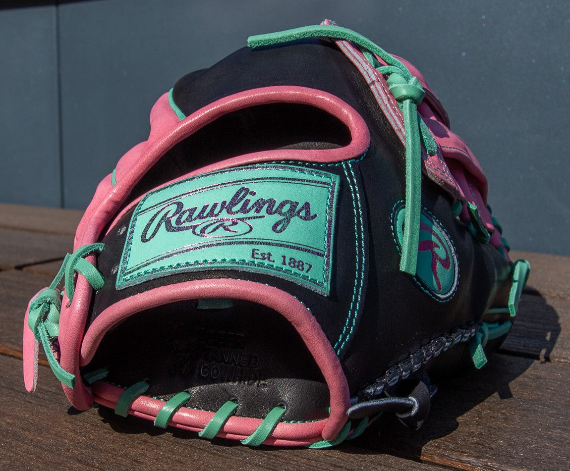A close-up of the Rawlings Heart of the Hide 12.25" Color Sync Baseball Glove (PROR207-6BP) in black, pink, and teal rests on wood, showcasing detailed stitching and prominent Rawlings branding.