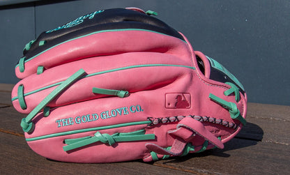 A Rawlings Heart of the Hide 12.25" Color Sync Baseball Glove (PROR207-6BP) in pink and teal, featuring THE GOLD GLOVE CO. embroidery, rests on a wooden surface against a dark background.