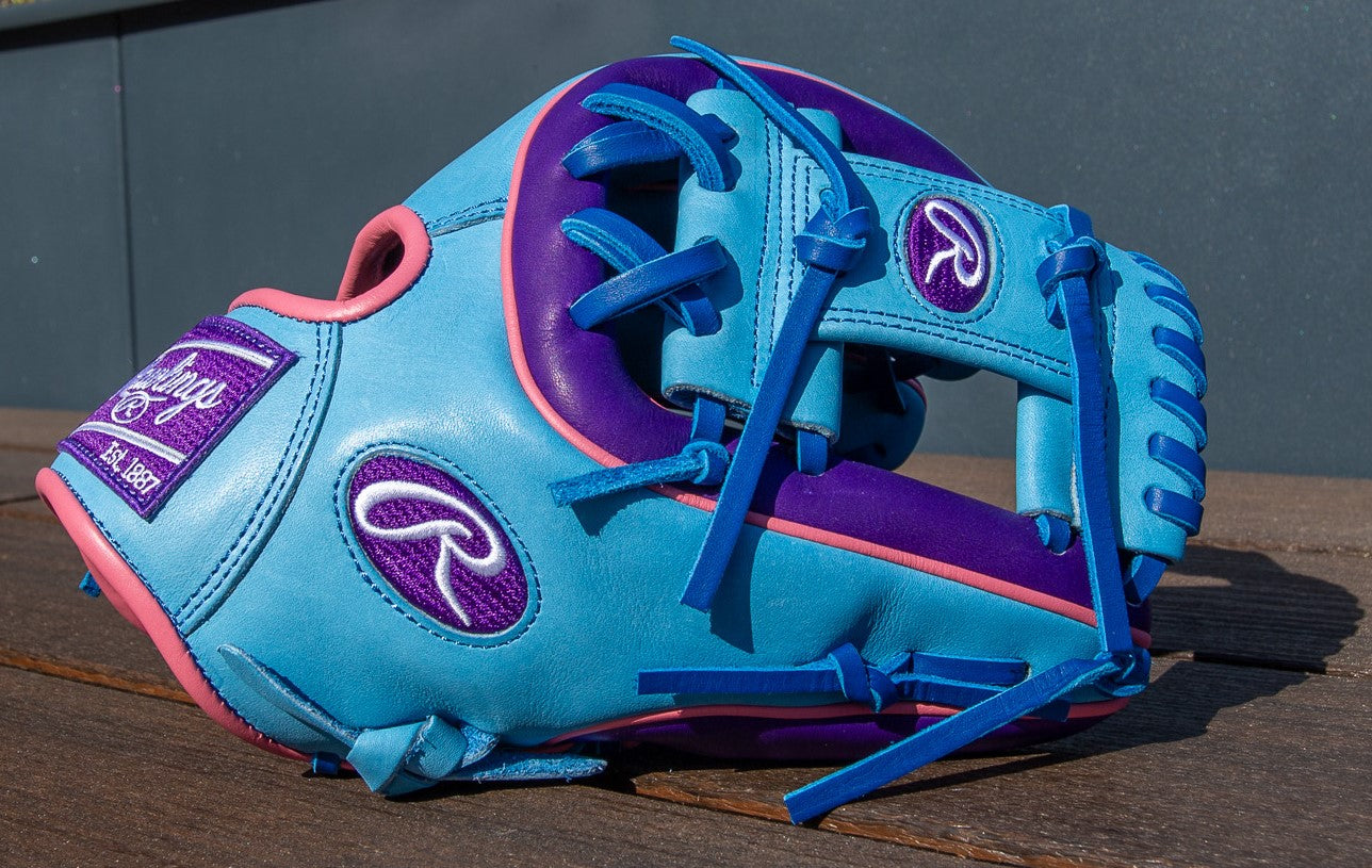 A Rawlings Heart of the Hide 11.5" Color Sync Baseball Glove (PROR314-2CBPU), blue with purple and pink accents, blue laces, purple interior details, and prominent Rawlings logos rests on a wooden surface.