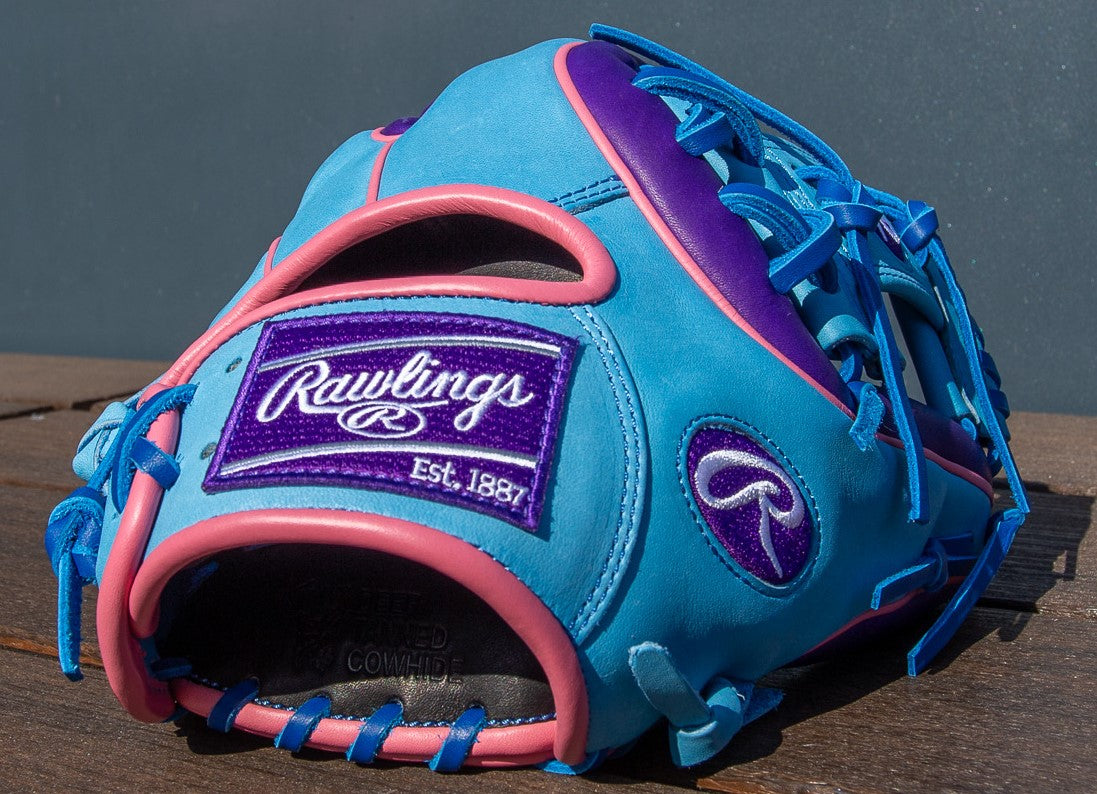 A blue Rawlings Heart of the Hide 11.5" Color Sync Baseball Glove (PROR314-2CBPU) with pink and purple accents, plus a purple patch embroidered with the Rawlings logo and "Est. 1887," rests on a wooden surface.