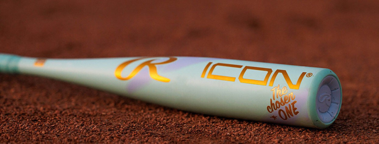 A close-up of a mint green Rawlings ICON baseball bat with gold lettering that reads ICON and The Chosen ONE, lying on a reddish-brown dirt surface.