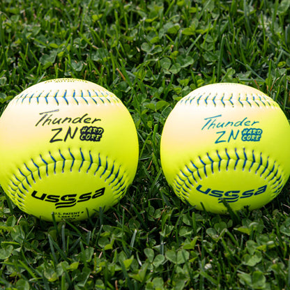Two Dudley USSSA Thunder ZN Hard Core PRO M 12" 44/375 Composite Slowpitch Softballs (model 4U12H) with clear labels rest on green grass, showcasing the quality Dudley is known for.