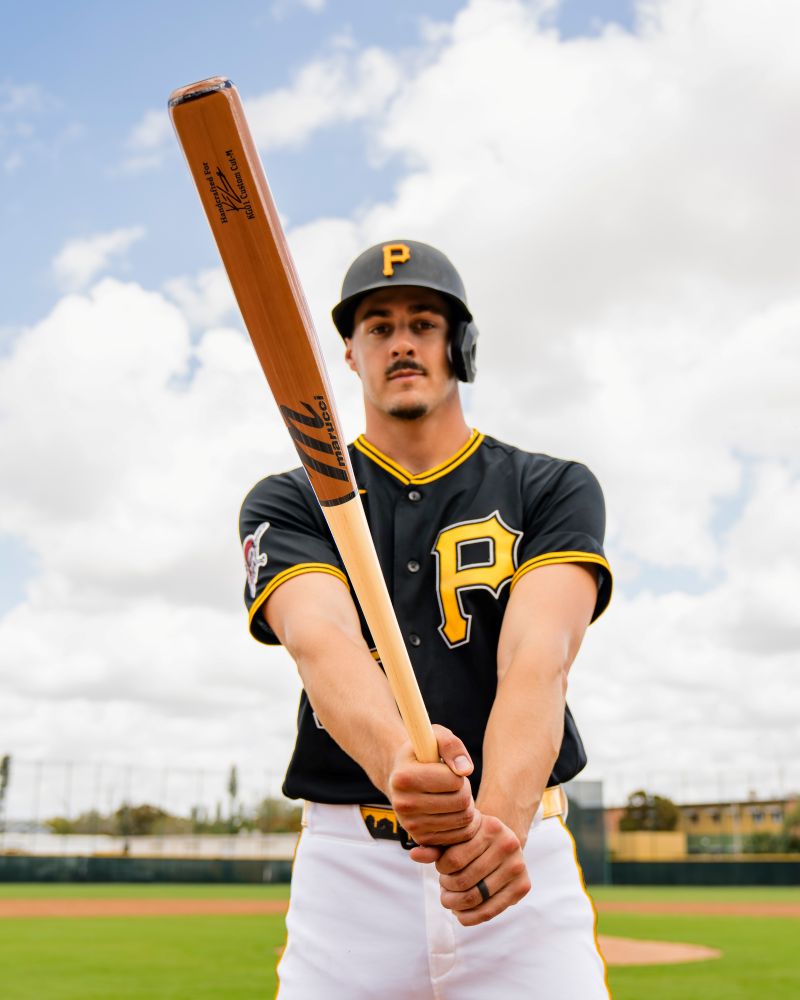 A baseball player in a black jersey with a yellow P holds a Marucci Konnor Griffin KG01 Pro Exclusive Maple Wood Bat (MVE6KG01) with a pro-x finish toward the camera on a baseball field under a partly cloudy sky.