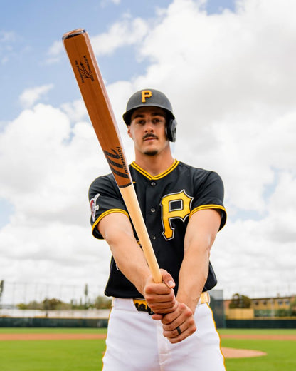 A baseball player in a black jersey with a yellow P holds a Marucci Konnor Griffin KG01 Pro Exclusive Maple Wood Bat (MVE6KG01) with a pro-x finish toward the camera on a baseball field under a partly cloudy sky.