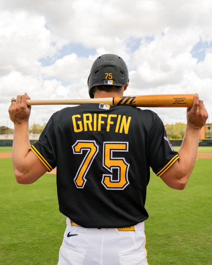 A baseball player wearing a black jersey with GRIFFIN and the number 75 stands on a field facing away from the camera, holding a Marucci Konnor Griffin KG01 Pro Exclusive Maple Wood Bat: MVE6KG01 with a pro-x finish behind his shoulders.
