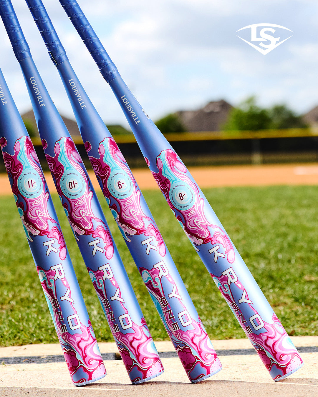 Four vibrant 2027 Louisville Slugger Kryo One (-10) Fastpitch Softball Bats (model WBL4273010), featuring a striking blue and pink swirl design, are lined up vertically on the grass near the ballfield. Each one-piece composite bat from Louisville Slugger stands ready for action, with the background showcasing a softly blurred fence and houses.