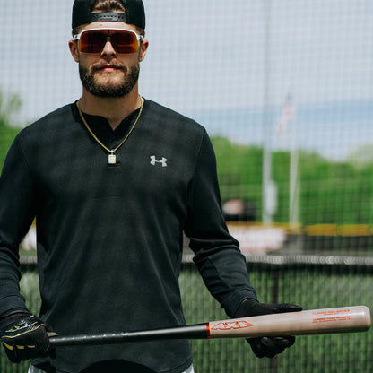 A baseball player stands on a field holding the AXE Flared Pro Series Maple Wood Baseball Bat: L124K-FLR, wearing sunglasses, a black Under Armour shirt, gloves, and a gold necklace, with a fence and trees in the background.