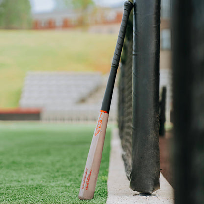 An AXE Flared Pro Series Maple Wood Baseball Bat (L124K-FLR) with a black handle stands upright against a black padded fence on the field, with bleachers and green grass seen in the background.