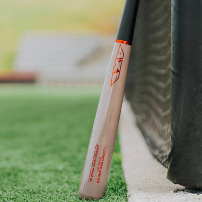 The AXE Flared Pro Series Maple Wood Baseball Bat: L124K-FLR, featuring red AXE branding and a pro flared handle, leans against a black net on a grassy baseball field with blurred stadium seating in the background.