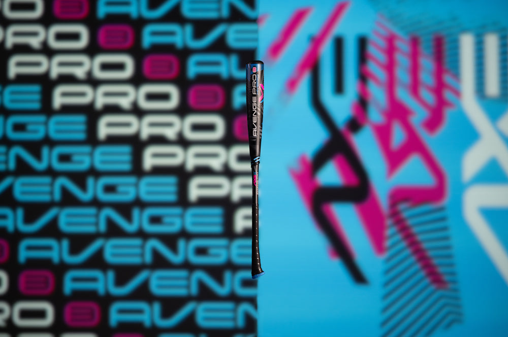 A youth bat stands upright between a black AVENGE PRO-patterned side and a blue side with black and pink designs—showcasing the 2024 AXE Avenge Pro 3 Hybrid (-8) USA Baseball Bat: L174M.