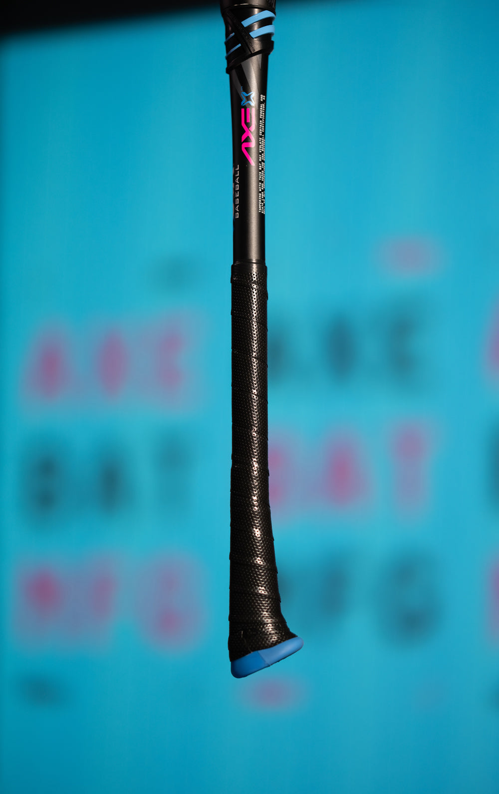 A close-up of the 2024 AXE Avenge Pro 3 Hybrid (-8) USA Baseball Bat handle, black with blue and pink accents, set against a blurred blue background and featuring indistinct pink and black lettering.