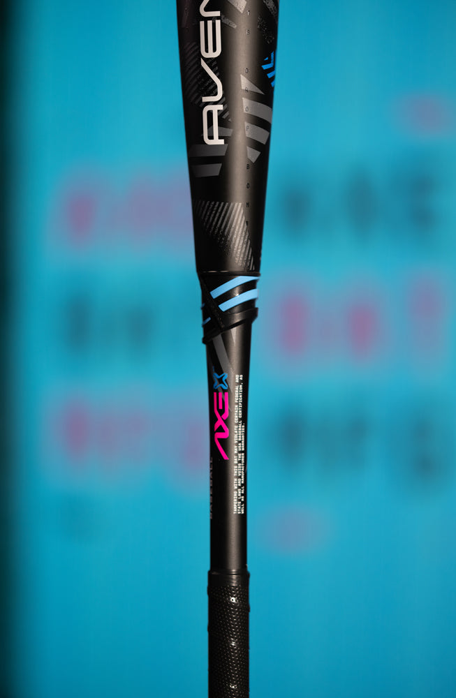 Close-up of the 2024 AXE Avenge Pro 3 Hybrid (-8) 2 5/8" USA Baseball Bat (L174M) in black with silver, blue, and pink accents; AXE branding stands out against a blurred blue background, highlighting its advanced design.