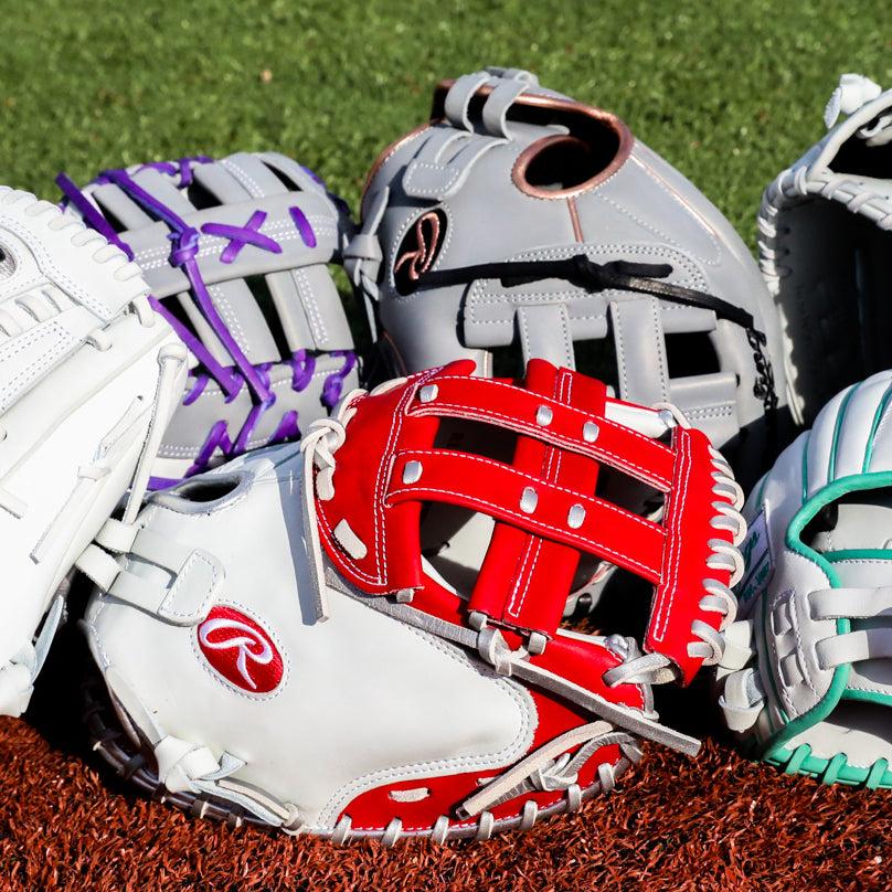 A close-up of five colorful baseball gloves, including white, gray, red, and purple designs, arranged on a brown turf surface with green grass in the background.