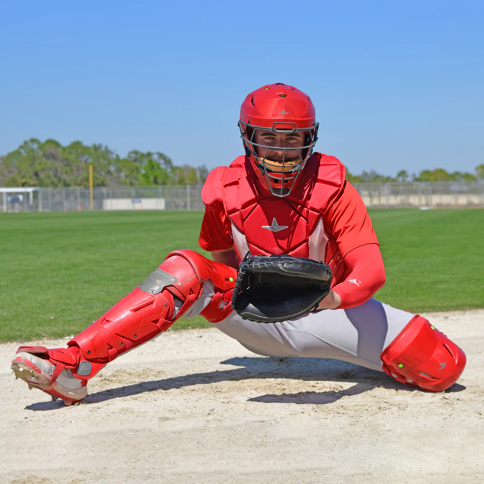 Wearing All Star MVP Pro Catcher's Leg Guards (LG-5) in red, the catcher crouches on the dirt field with glove outstretched, ready for a pitch. Green grass, trees, and blue sky fill the background.