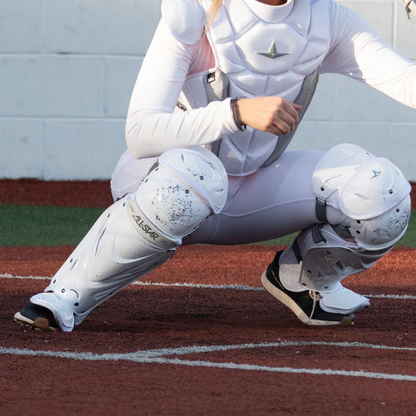 An All Star PHX Paige Halstead Fastpitch catcher in white protective gear crouches behind home plate, wearing All Star LGW-PHX leg guards with the LINQ hinge system for flexibility and Delta-Flex calf harness for a secure fit.