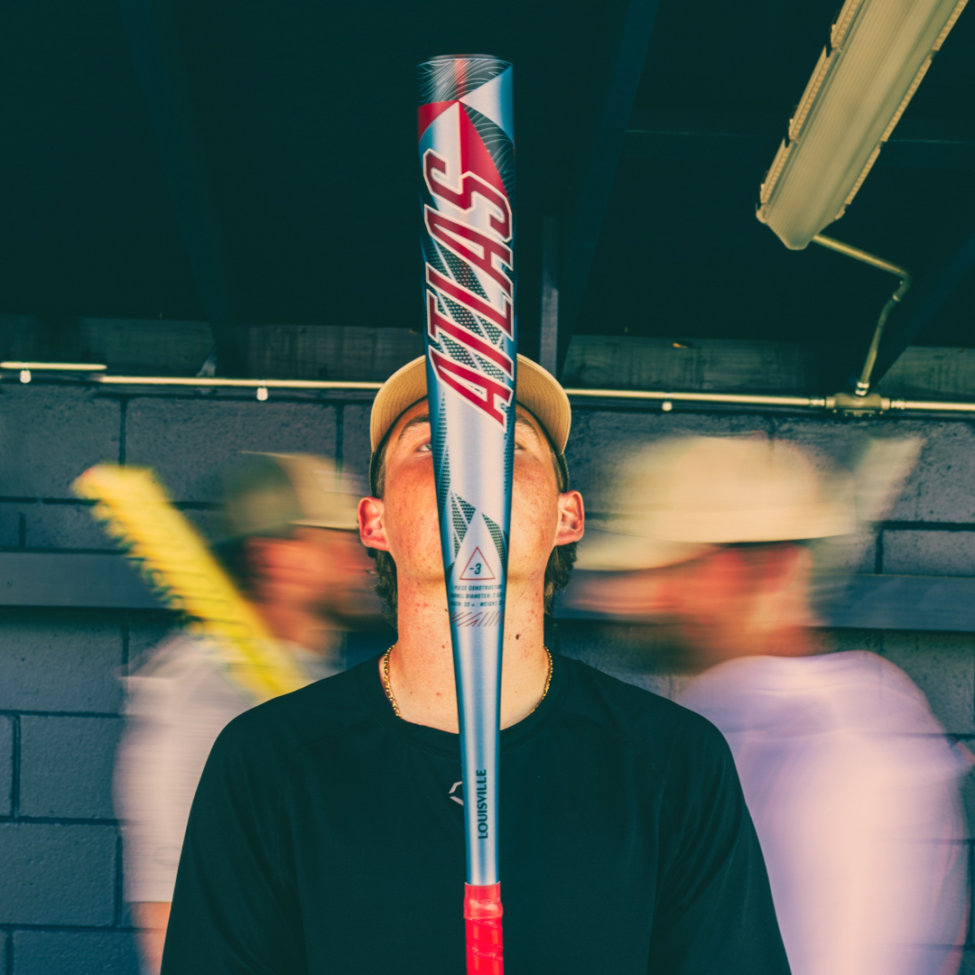 Someone balances a 2026 Louisville Slugger Atlas (-3) BBCOR Baseball Bat (WBL4116010) on their face, with blurred figures in the background against a blue wall.