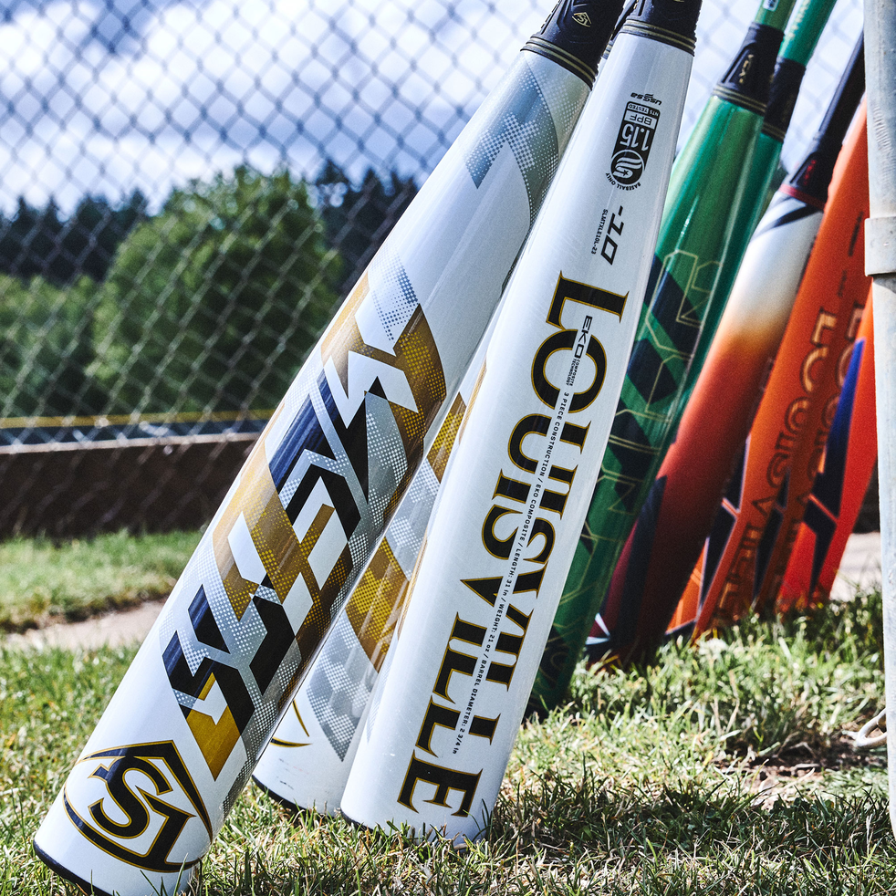 Several colorful baseball bats, including a prominent white and gold Louisville Slugger bat, are lined up on grass next to a chain-link fence on a sunny day. Trees and sky are visible in the background.