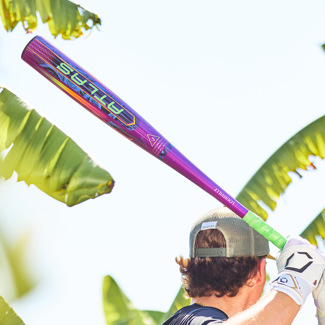 A person in a cap and batting gloves holds the 2025 Louisville Slugger Atlas Neon Energy (-3) BBCOR Baseball Bat (WBL4164010), poised to swing, with large green banana leaves and bright sunlight in the background.