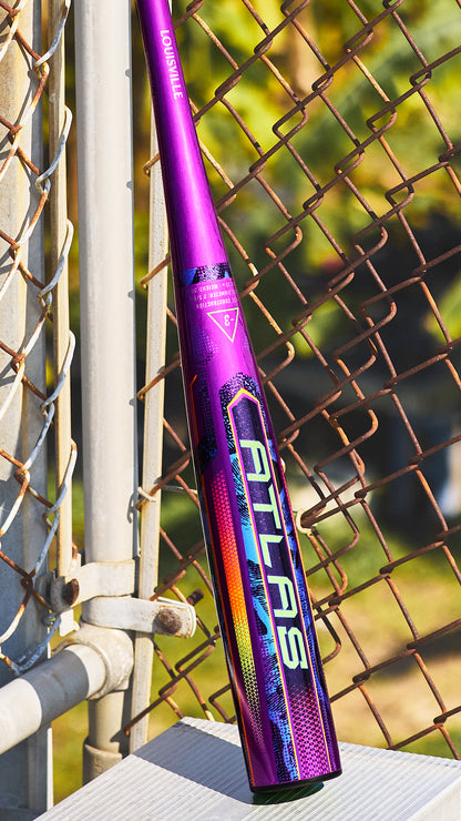 A 2025 Louisville Slugger Atlas Neon Energy (-3) BBCOR Baseball Bat (WBL4164010) rests against a metal fence at a baseball field, sunlight casting shadows while greenery fills the background.