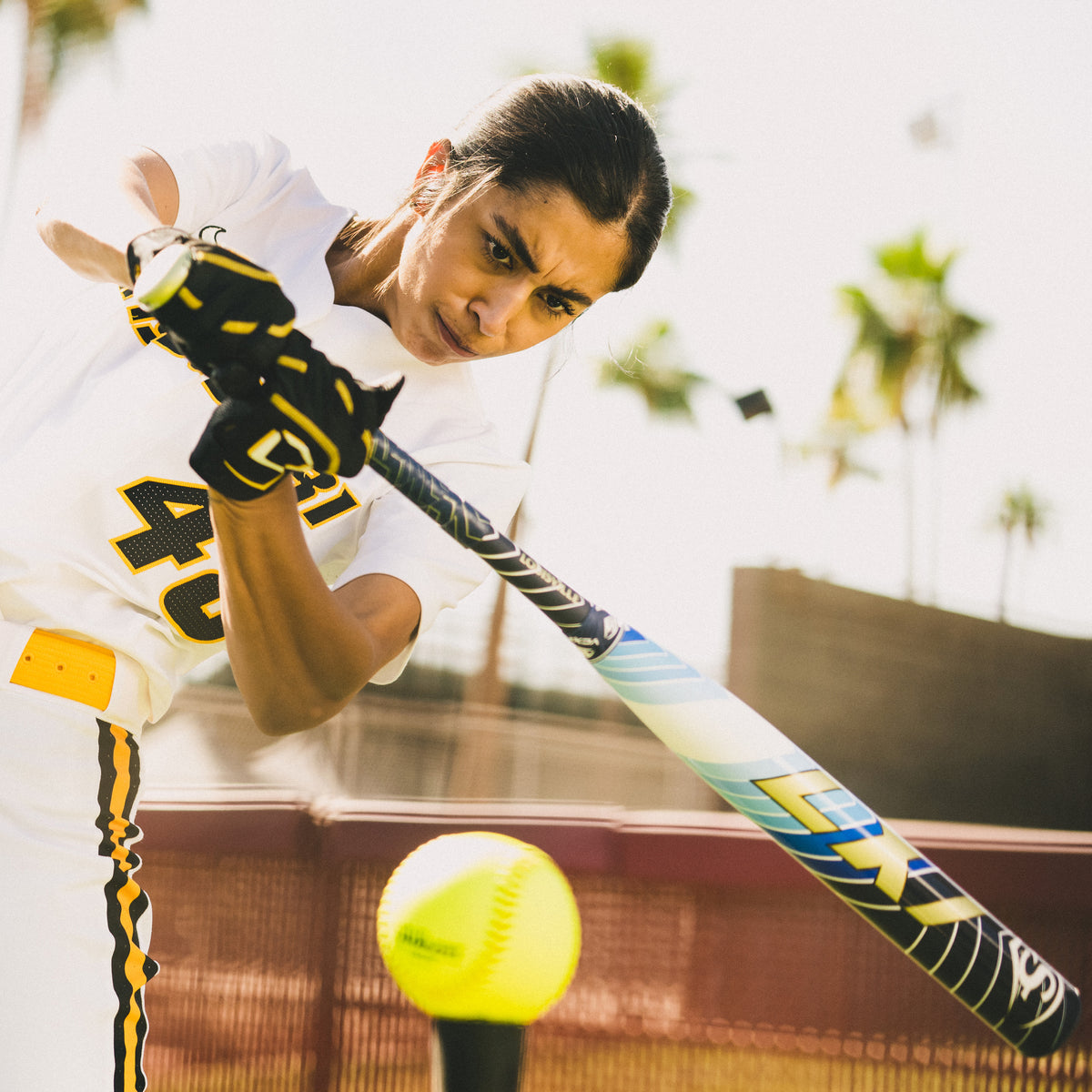 A focused softball player in a white and yellow uniform swings a bat at an incoming yellow softball during a sunny outdoor game, with palm trees and a fence in the background.