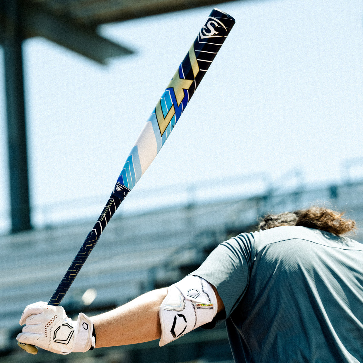 A baseball player with long hair, wearing a green shirt, white gloves, and an elbow guard, holds a colorful bat over their shoulder in an outdoor stadium. The background shows blurred bleachers and bright sunlight.