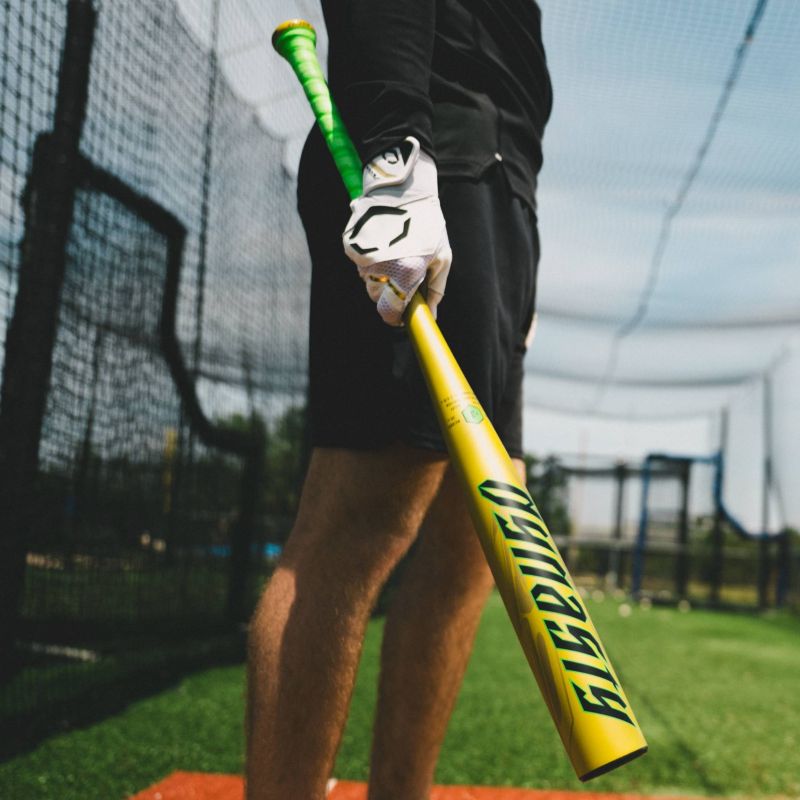 A person in black shorts and a white glove holds a bright yellow-green 2026 Louisville Slugger Dynasty (-3) BBCOR Baseball Bat (WBL4162010) inside a batting cage, standing on a red mat with nets and blue sky in the background.