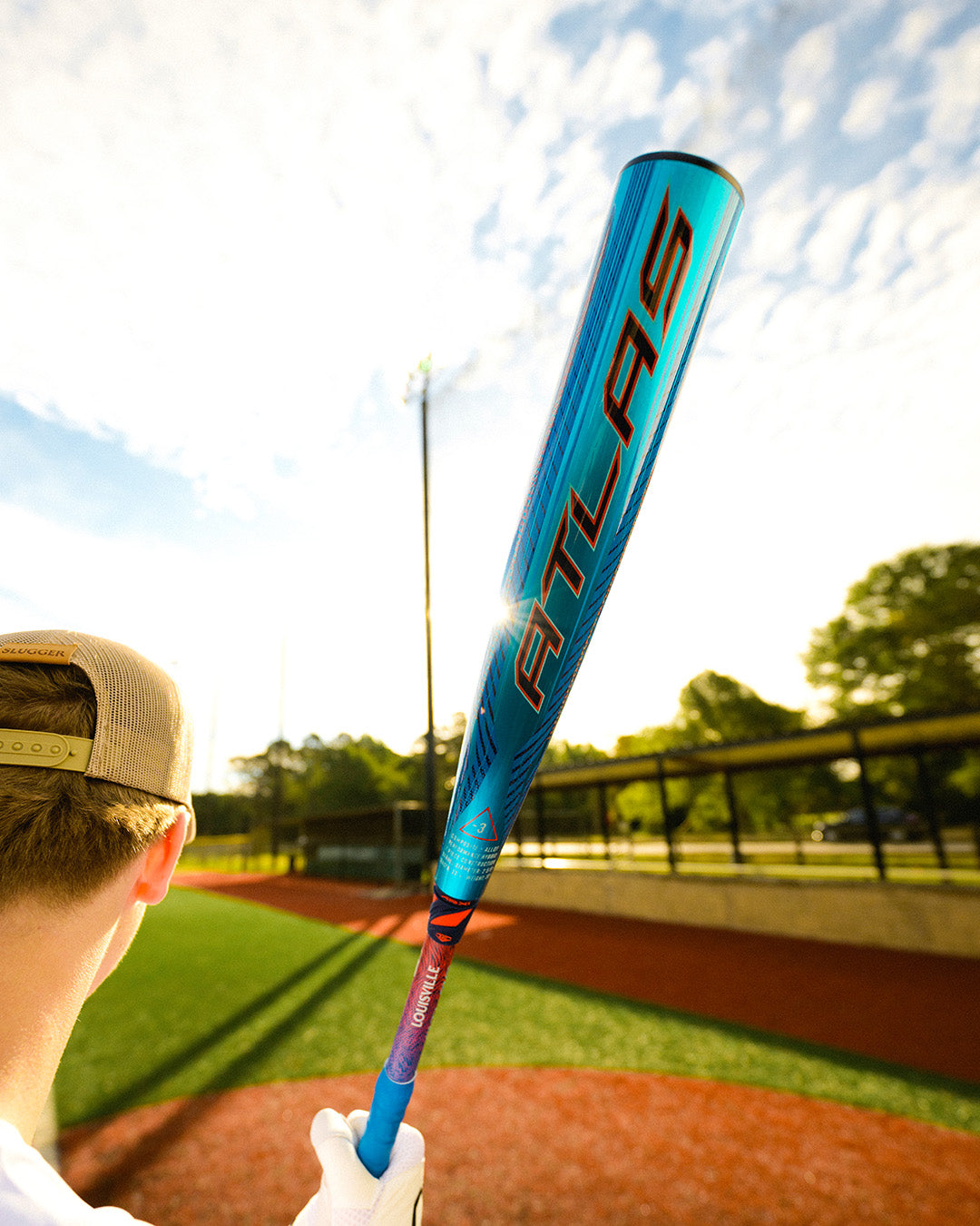 A person in a tan cap holds up the 2027 Louisville Slugger Atlas Hybrid (-3) BBCOR Baseball Bat (WBL4246010) with an EVOKE Alloy Barrel on a sunny day at an outdoor baseball field with green grass and red dirt.