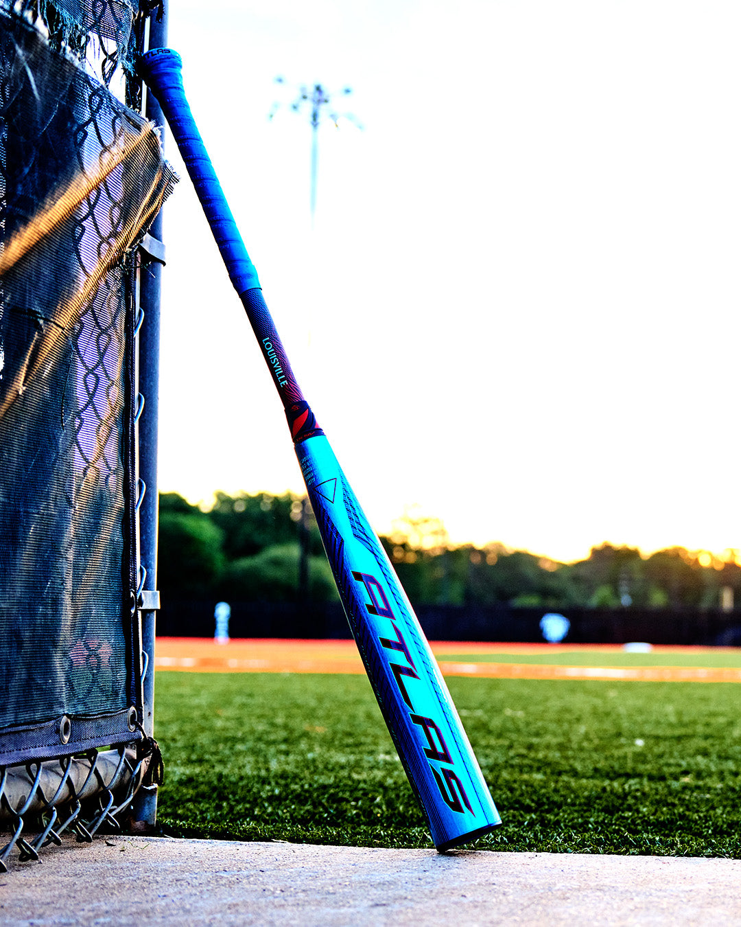 A 2027 Louisville Slugger Atlas Hybrid (-3) BBCOR Baseball Bat (WBL4246010) leans against a chain-link fence by a baseball field, with players and green trees blurred in the sunset background.