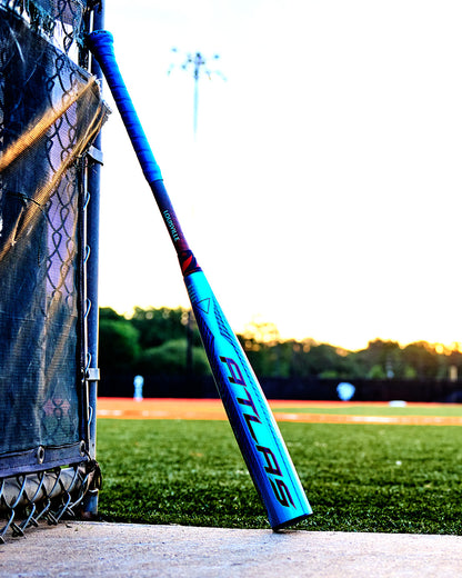 A 2027 Louisville Slugger Atlas Hybrid (-3) BBCOR Baseball Bat (WBL4246010) leans against a chain-link fence by a baseball field, with players and green trees blurred in the sunset background.