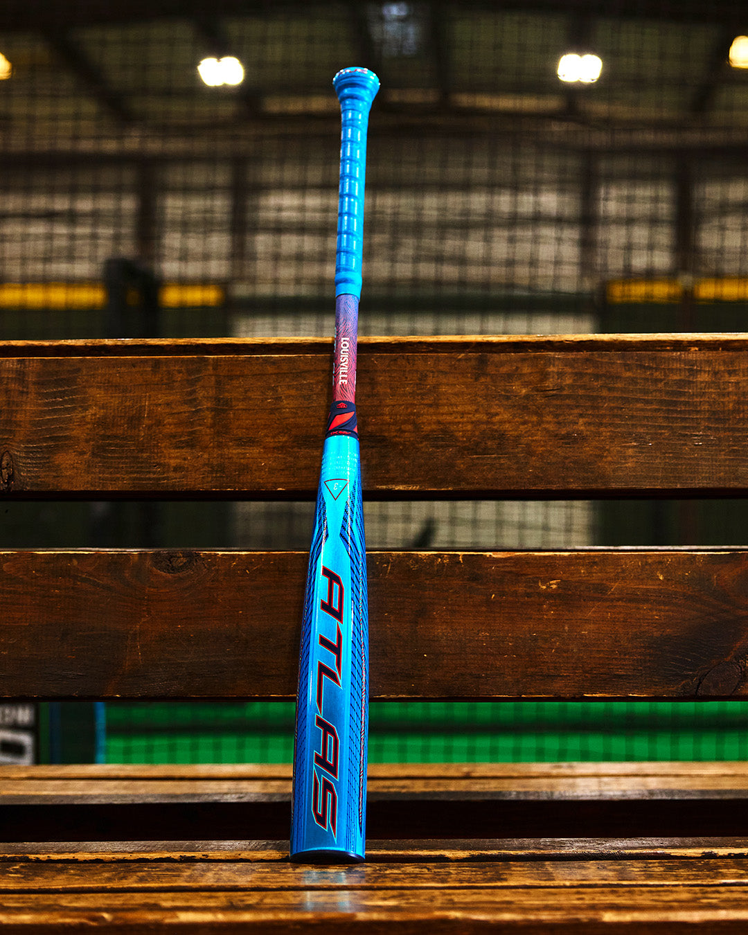 A blue and red Louisville Slugger 2027 Atlas Hybrid (-3) BBCOR Baseball Bat (WBL4246010) stands upright on a wooden bench in an indoor sports facility with netting and lights in the background.