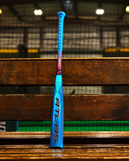 A blue and red Louisville Slugger 2027 Atlas Hybrid (-3) BBCOR Baseball Bat (WBL4246010) stands upright on a wooden bench in an indoor sports facility with netting and lights in the background.