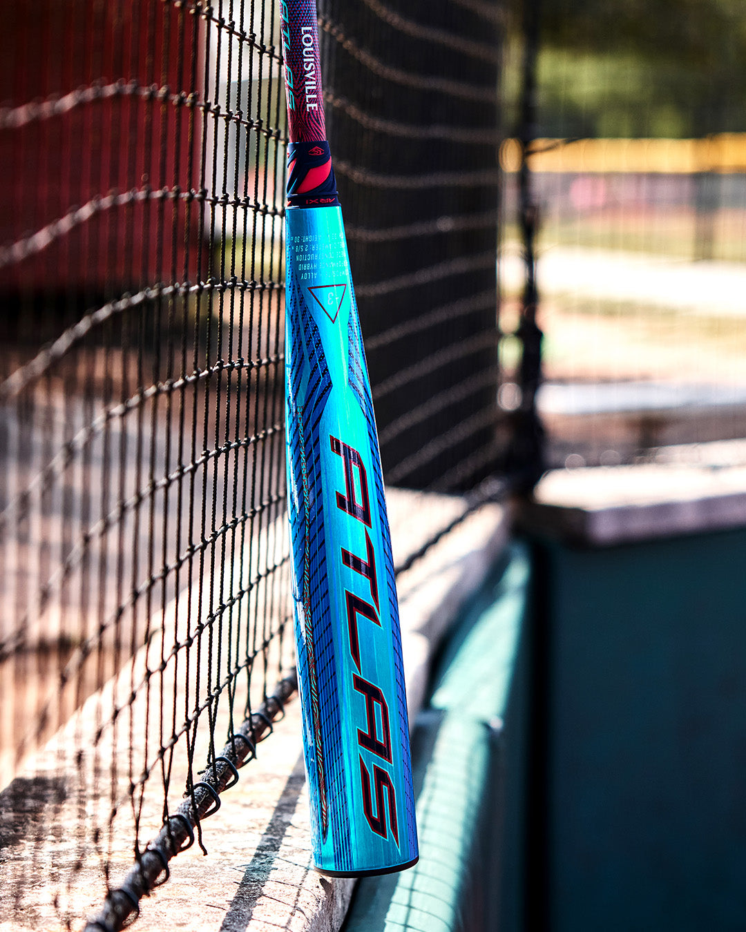 A 2027 Louisville Slugger Atlas Hybrid (-3) BBCOR Baseball Bat (WBL4246010), featuring "ATLAS" lettering, leans against a fence at a sunny outdoor baseball field.