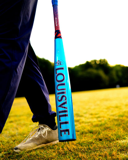 A person in white shoes and dark pants stands on grass, holding a blue Louisville Slugger Atlas Hybrid (-3) BBCOR Baseball Bat (WBL4246010), with sunlit trees in the background.