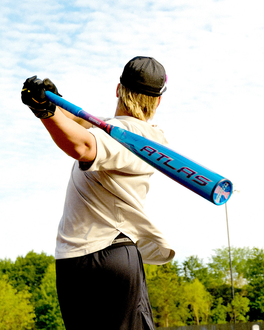 A person in a cap and glove holds a blue 2027 Louisville Slugger Atlas Hybrid (-3) BBCOR Baseball Bat (WBL4246010) over their shoulder outdoors, with trees and a cloudy sky behind them.