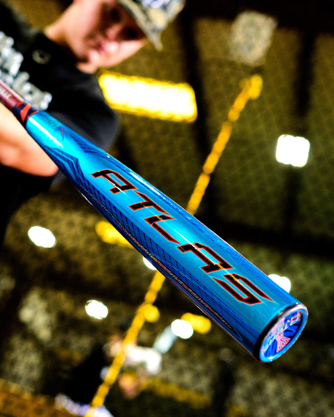 A person holds the 2027 Louisville Slugger Atlas Hybrid (-3) BBCOR Baseball Bat (WBL4246010), showcasing the blue EVOKE Alloy Barrel and red ATLAS letters, with an indoor batting cage blurred in the background.