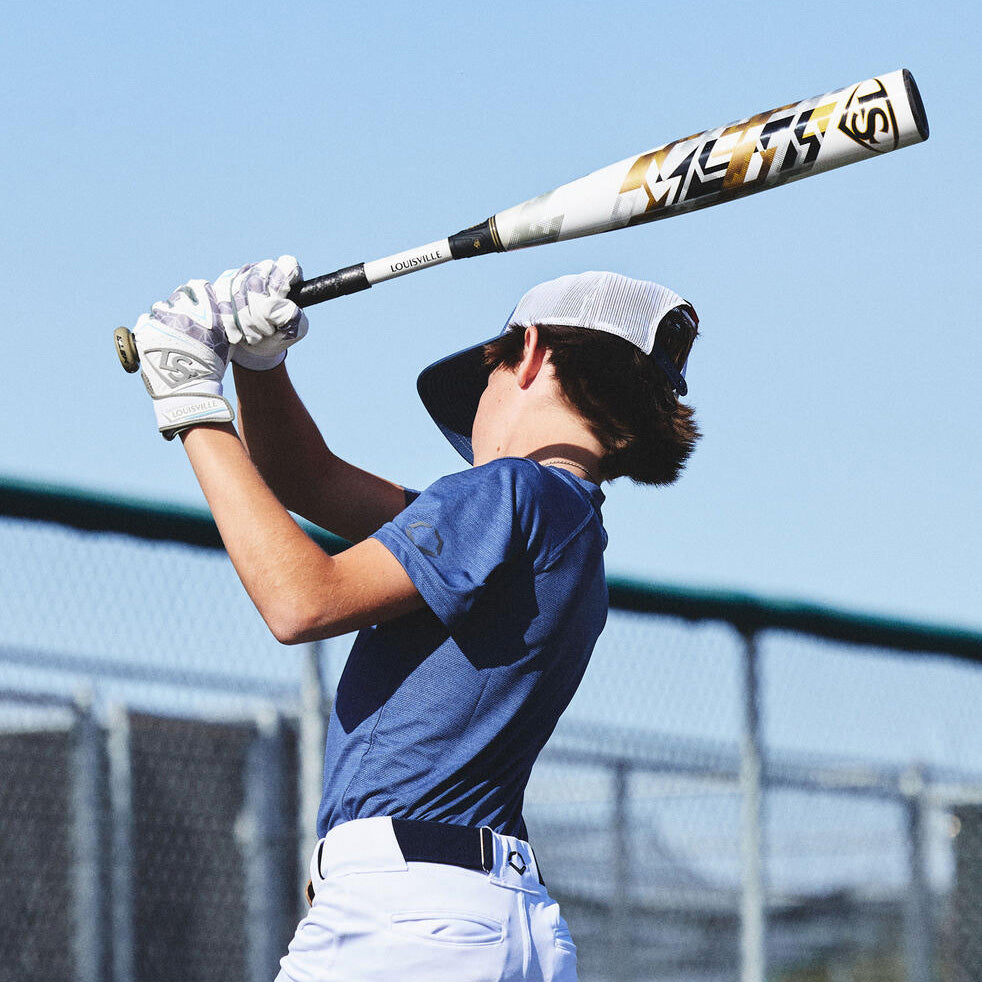 A young baseball player in a blue shirt and white pants swings a bat, wearing a white cap and gloves. He stands near a chain-link fence under a clear blue sky.
