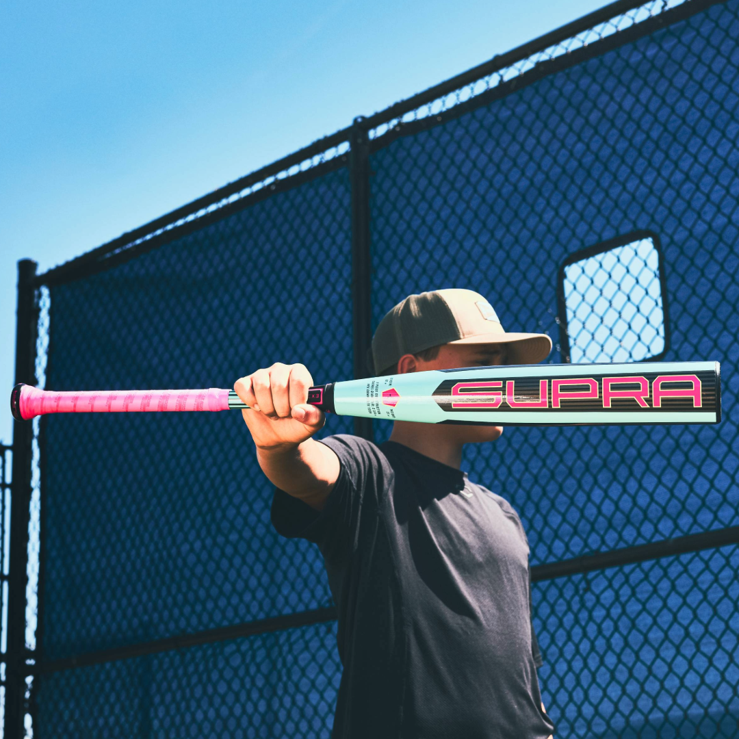 A person in a cap and dark shirt holds the 2026 Louisville Slugger Supra (-5) USSSA Baseball Bat (WBL4120010) outstretched, standing before a blue chain-link fence under a clear sky.