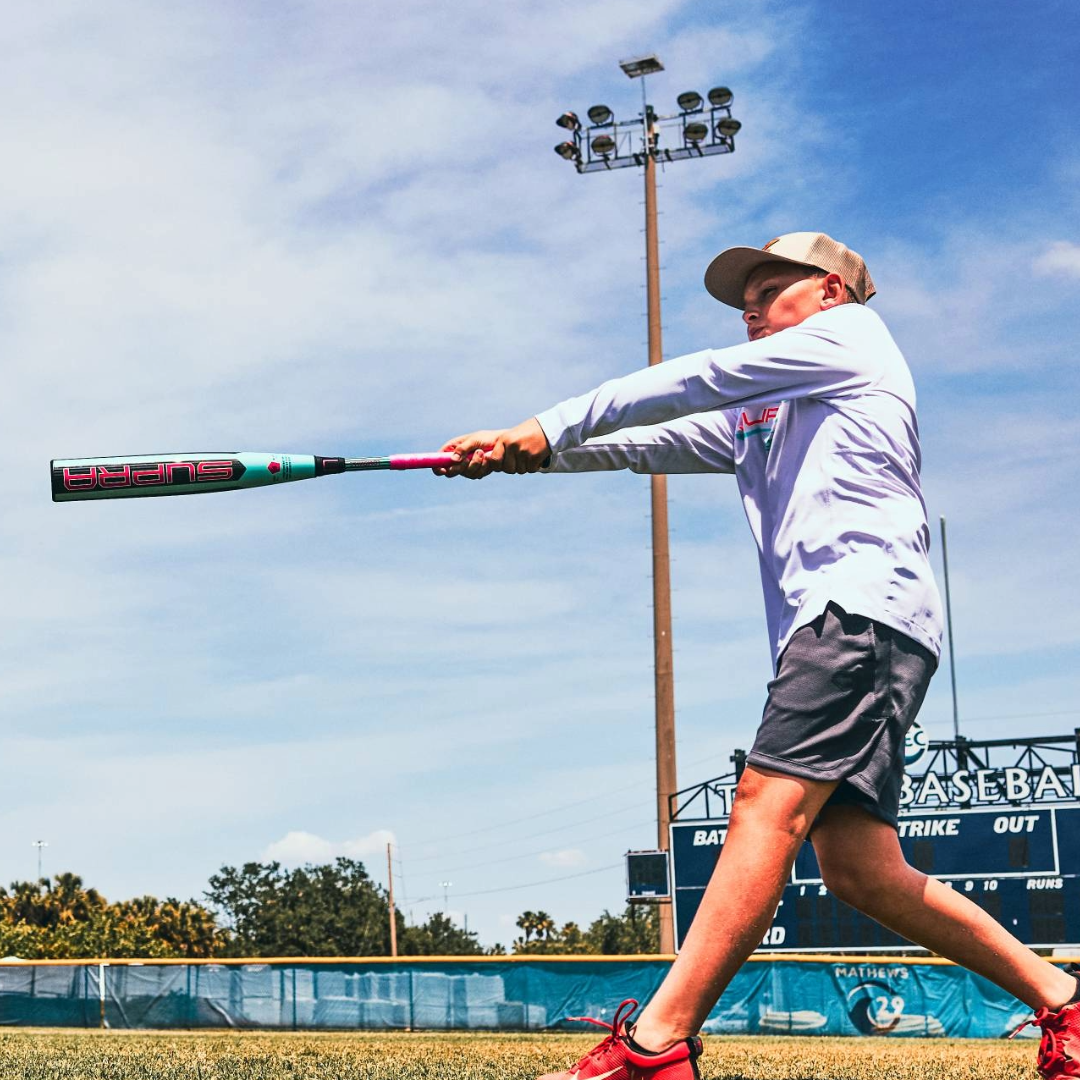 A young boy in a white shirt, gray shorts, and a cap swings the 2026 Louisville Slugger Supra (-5) 2 3/4" USSSA Baseball Bat (WBL4120010) on a sunny field with a scoreboard and tall lights in the background.