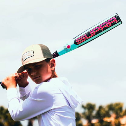 A boy in a cap and white shirt holds the 2026 Louisville Slugger Supra (-5) USSSA Baseball Bat (WBL4120010) over his shoulder, ready to swing—an ideal moment for travel ball, trees softly blurred in the background.