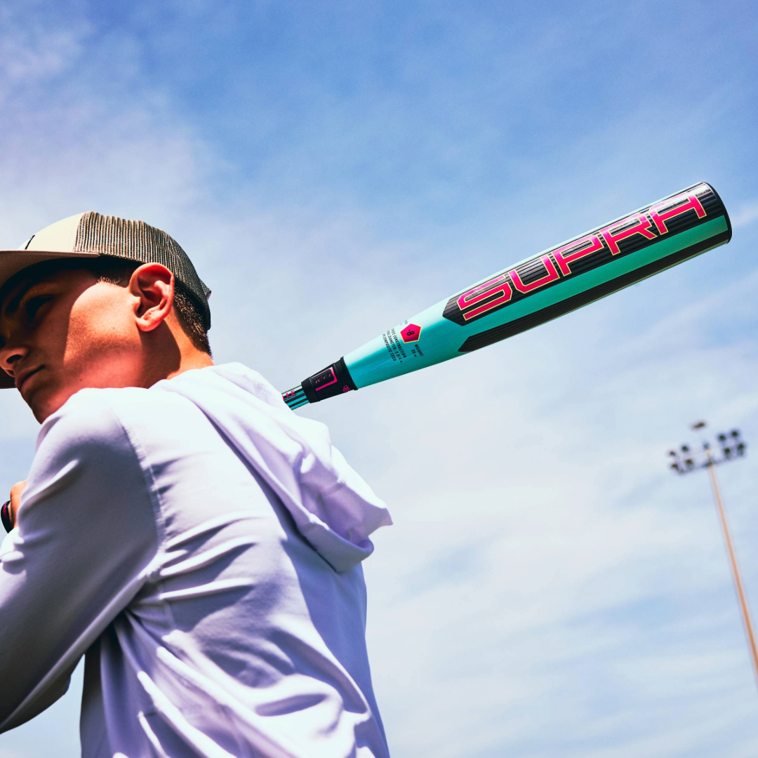 A young player in a white hoodie and cap holds the 2026 Louisville Slugger Supra (-5) USSSA Baseball Bat (WBL4120010) over their shoulder outdoors—ideal for travel ball athletes.